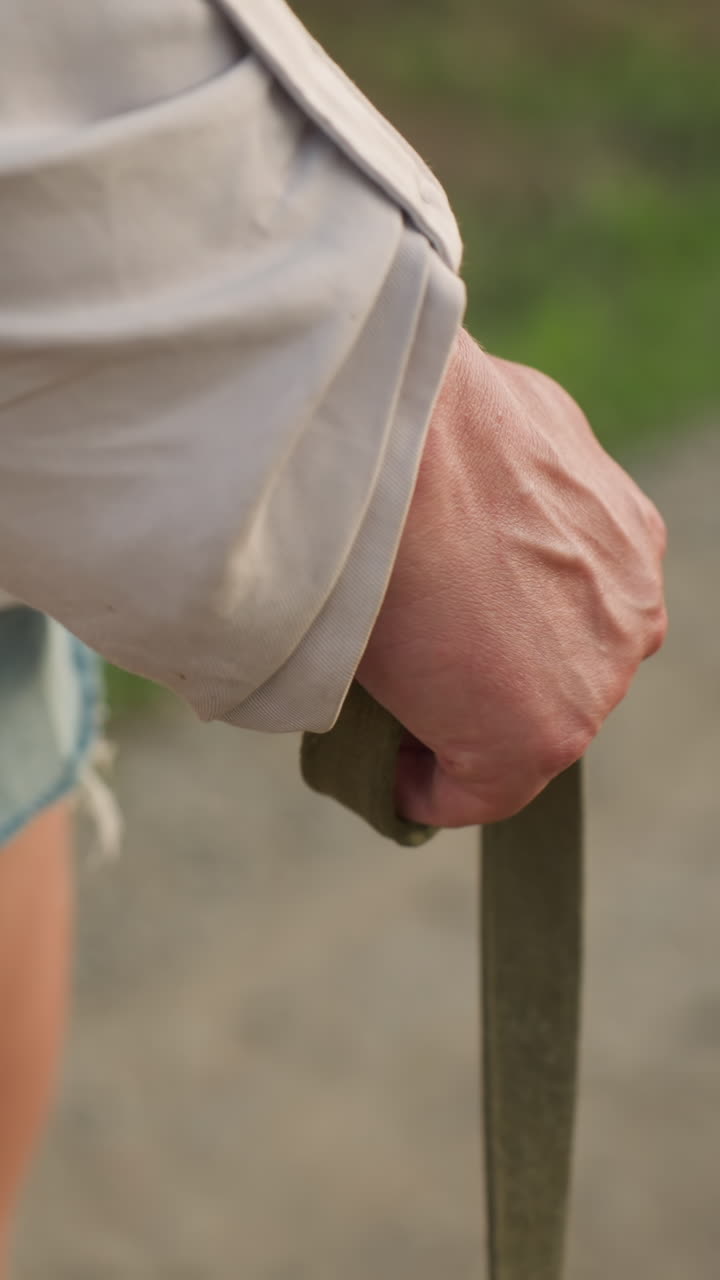 cerca de la mano sosteniendo la correa mientras camina por un camino de tierra, con ropa casual, incluidos pantalones cortos de jean, venas en la mano visibles, luz solar brillante iluminando la vegetación en un fondo borroso