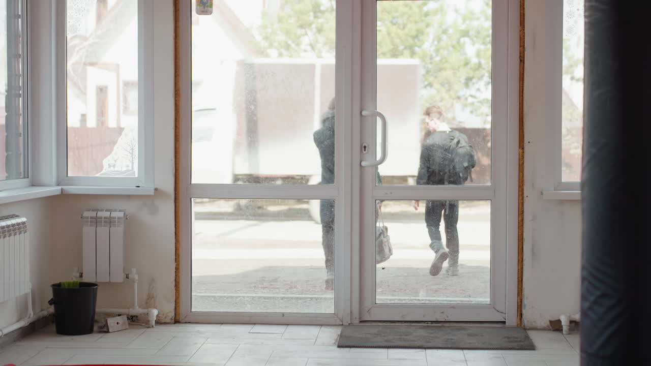 Two guys walking out of training hall, one holding large brown leather bag, both wearing dark clothing and boots, sunlight illuminating interior with red mats and radiators visible inside