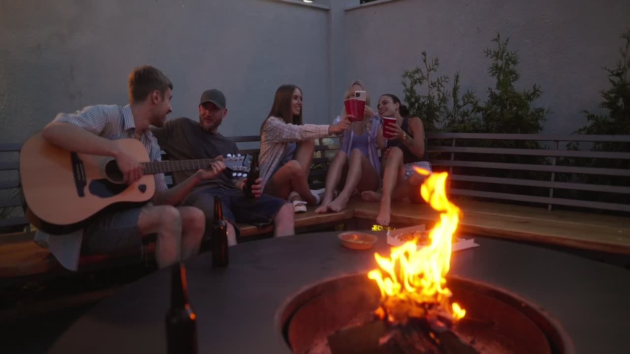 un grupo alegre de amigos cantan escuchan canciones en la guitarra y se comunican mientras pasan tiempo cerca de un fuego en el patio de una casa de campo