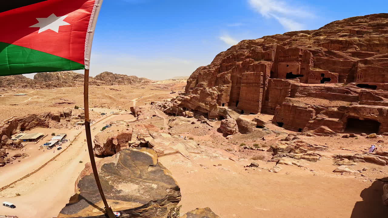 Approaching the flag of Hashemite Kingdom of Jordan waving in the wind. View on the ancient city of Petra from top of the rock.