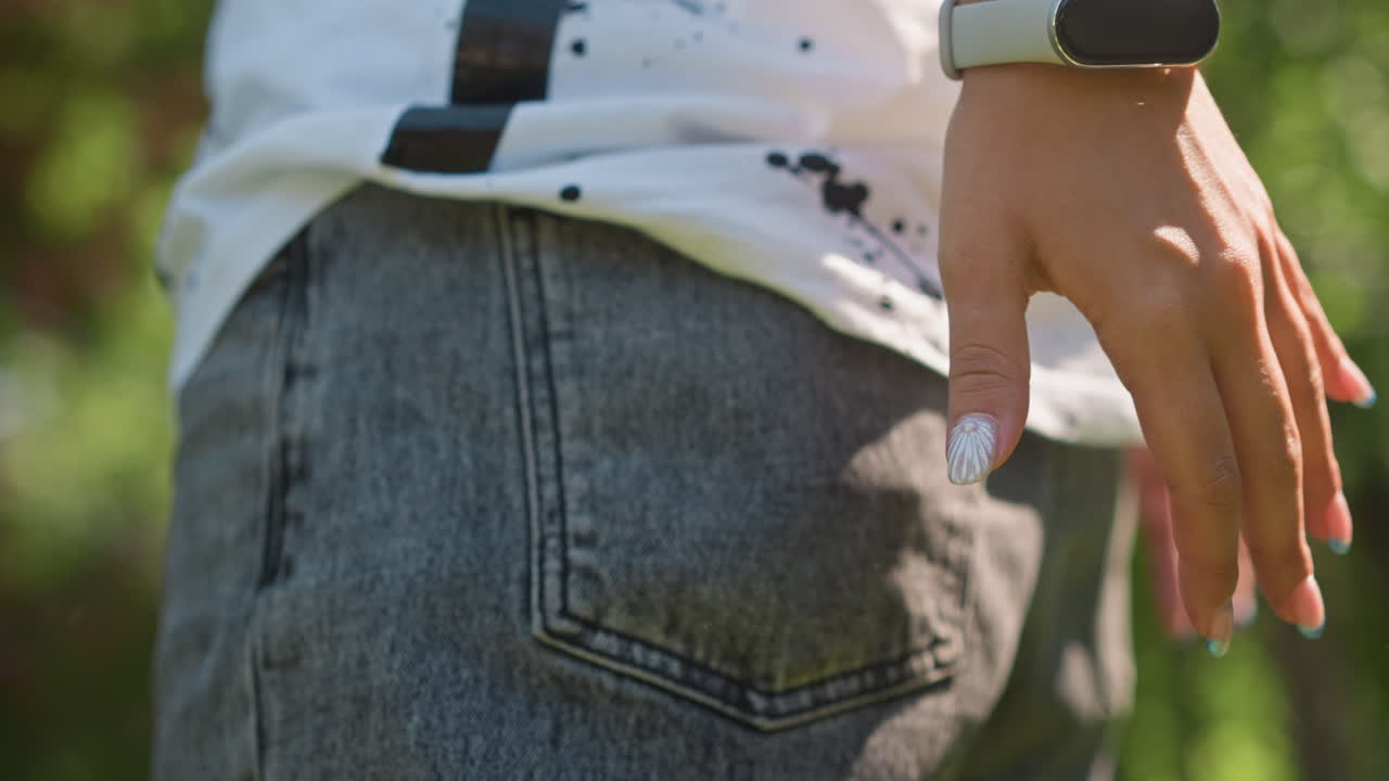 Närbild av en stänkt skjorta över en denimficka medan en hand glider ner i bakfickan. En streetskater kontrollerar omgivningen, smartklocka på handleden, solbelyst parkbakgrund, texturerade syratvättade jeans och avslappnad hållning