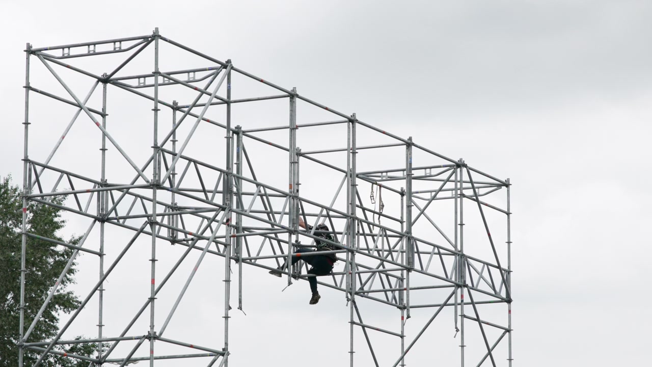 Construction worker crouched on modular steel scaffolding with safety harness and lanyard on horizontal braces and platform beneath under overcast worksite sky