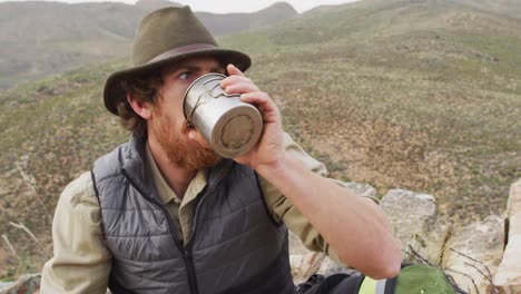 hombre caucásico barbudo sobreviviente sentado en la montaña en el desierto, bebiendo agua