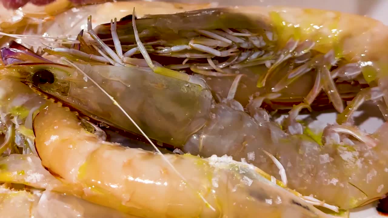 A hand sprinkles coarse salt over a close-up view of fresh prawns, highlighting their texture and preparation.