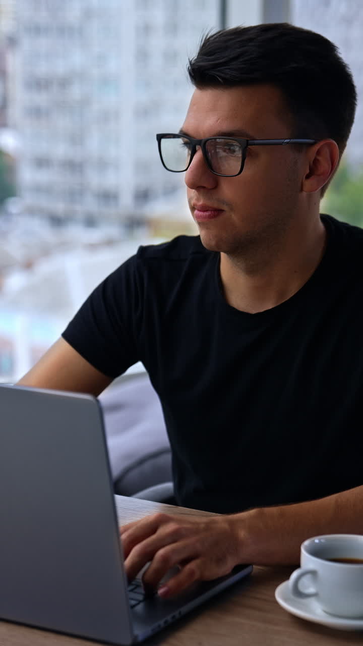 Pensive young man in glasses and black t-shirt sits in the office in front of laptop. Male looks into window thinking over something. Blurred backdrop. Vertical video