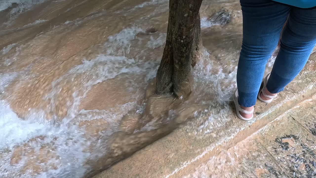 Individual in jeans and sneakers walks cautiously along a city sidewalk partially submerged by fast-moving floodwater, with natural daylight and handheld camera movement