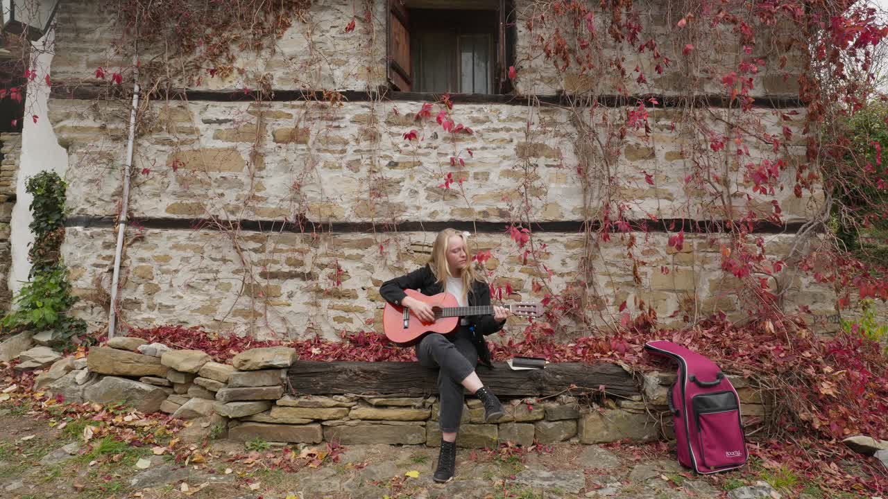 una joven compositora toca la guitarra, el follaje de otoño, la escena de la pared de piedra.