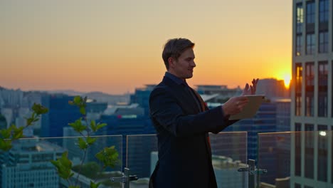in a good mood, a caucasian businessman has a video call on his tablet, showing the beautiful sunset and city skyline from a modern rooftop terrace
