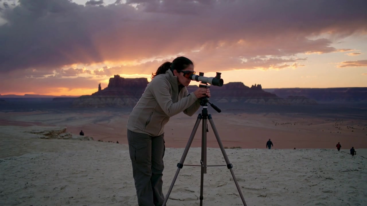 Al tramonto, una donna in pile regola un cannocchiale da osservazione su treppiede al bordo del deserto, scrutando le mesas