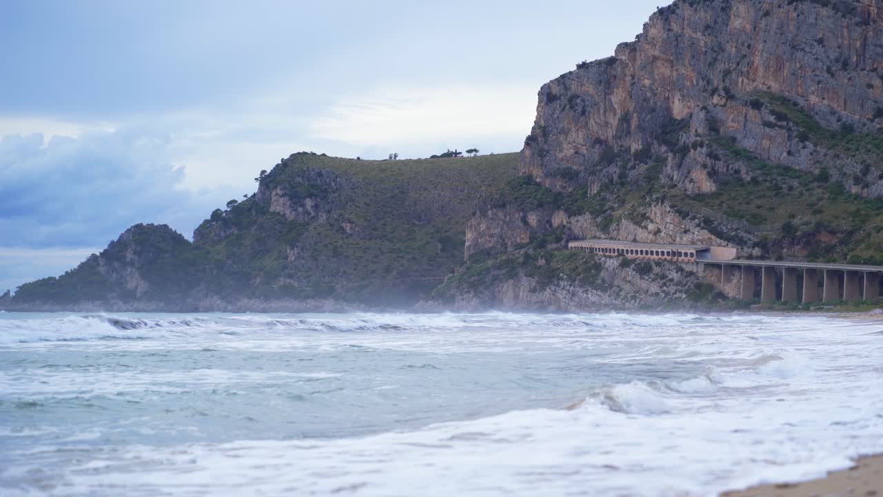 pintoresca vista a la playa de las olas blancas del océano rodando hacia y rompiendo en la playa de arena con el túnel del puente y la montaña en el fondo, gaeta, italia, estática