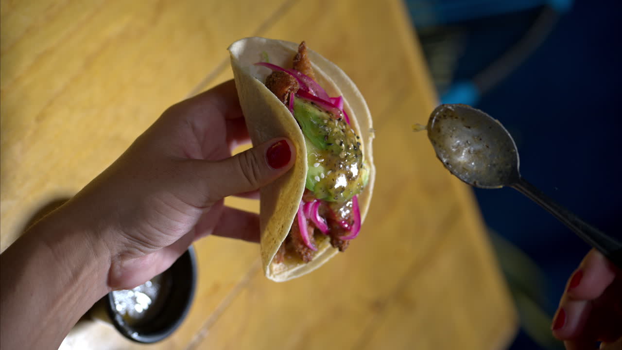 movimiento lento vertical de una mujer sosteniendo un taco de mariscos de camarón con cebolla roja y aguacate sirviendo vertiendo salsa verde en la parte superior en un restaurante tradicional mexicano