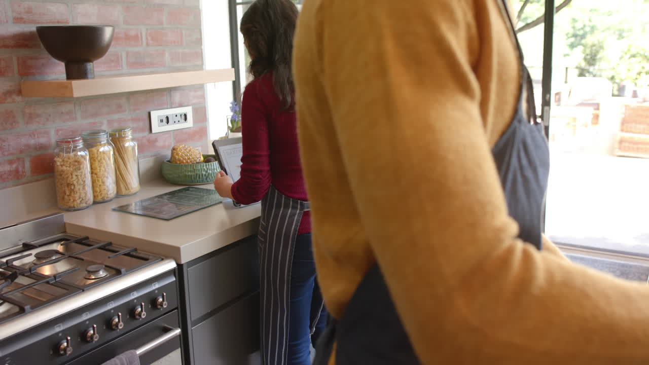 Coppia felice e diversificata con grembiuli che usa un tablet e cucina in una cucina soleggiata, al rallentatore