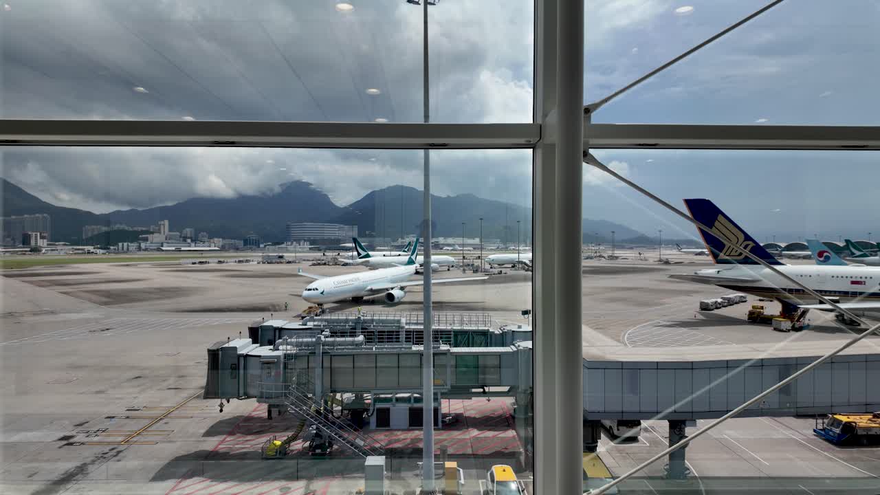 View of airplanes at Hong Kong airport through a glass structure. Features a jet bridge and runway, with a mountainous backdrop under a cloudy sky.