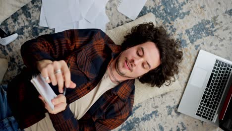 un tipo moreno pensativo con cabello rizado en una camisa a cuadros se acuesta en el suelo sobre una almohada y escribe sus ideas en un cuaderno blanco entre muchos papeles y una computadora portátil en casa