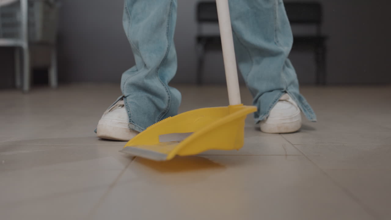Leg view of employer sweeping floor with broom and dustpan inside modern laundromat, stainless washers background, jeans and sneakers visible, collecting debris after filter service