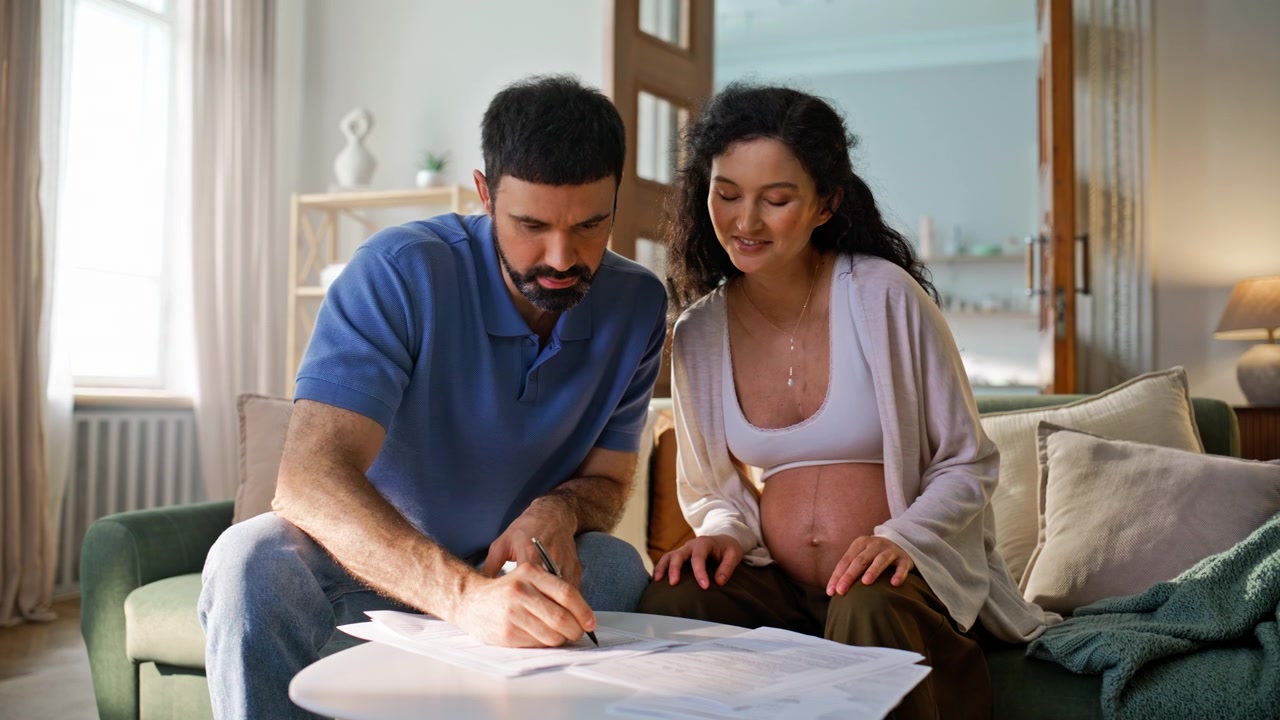 Famiglia che firma documenti sul divano di casa. Uomo che compila documenti accanto alla moglie incinta.