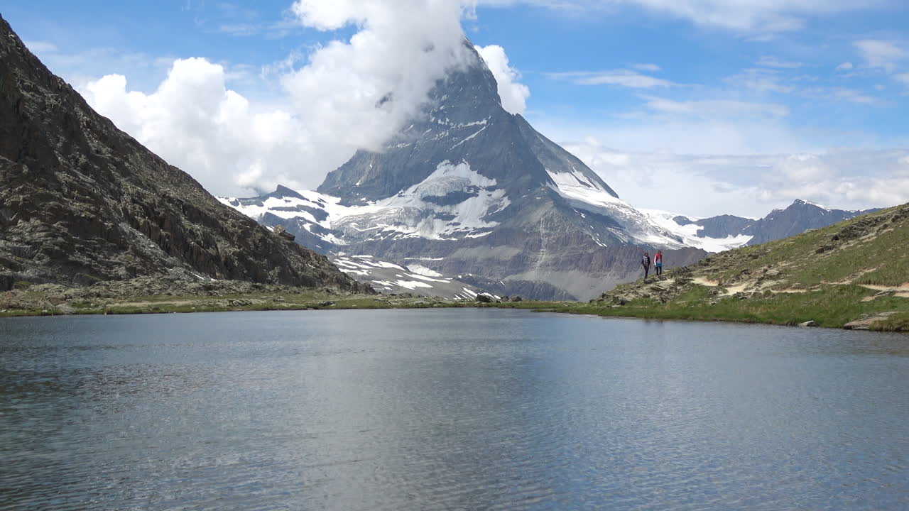 Veduta panoramica di un tranquillo lago alpino con la vetta del Cervino e ghiacciai sullo sfondo, escursionisti su una cresta, in una giornata di sole a Zermatt, Vallese, Svizzera