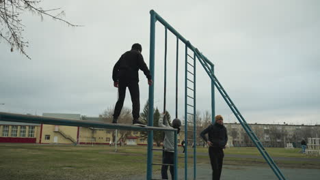 a young boy dressed in a black outfit carefully walks on an iron pole while another person descends the iron rail and walks away, meanwhile, a boy in a gray sweater climbs the iron structure