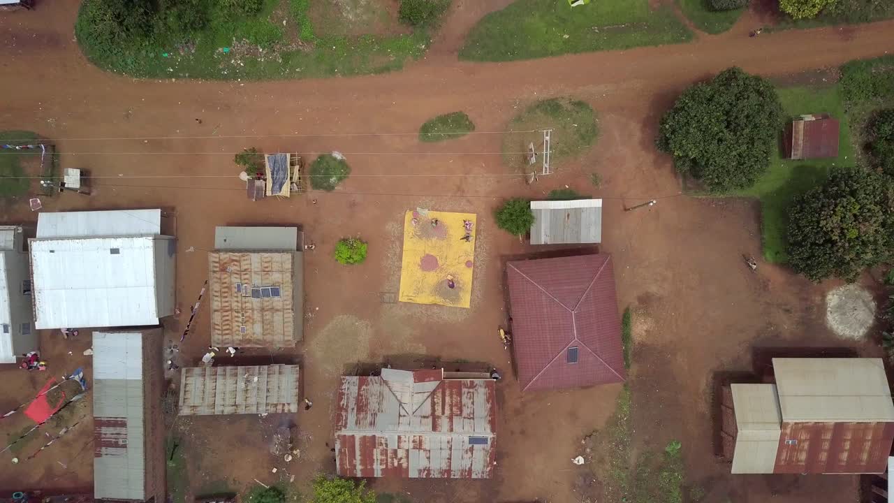 Ripresa aerea dall'alto di un piccolo insediamento rurale in Uganda, con semplici case dai tetti in metallo e un telo giallo centrale dove le persone essiccano chicchi di sorgo al sole