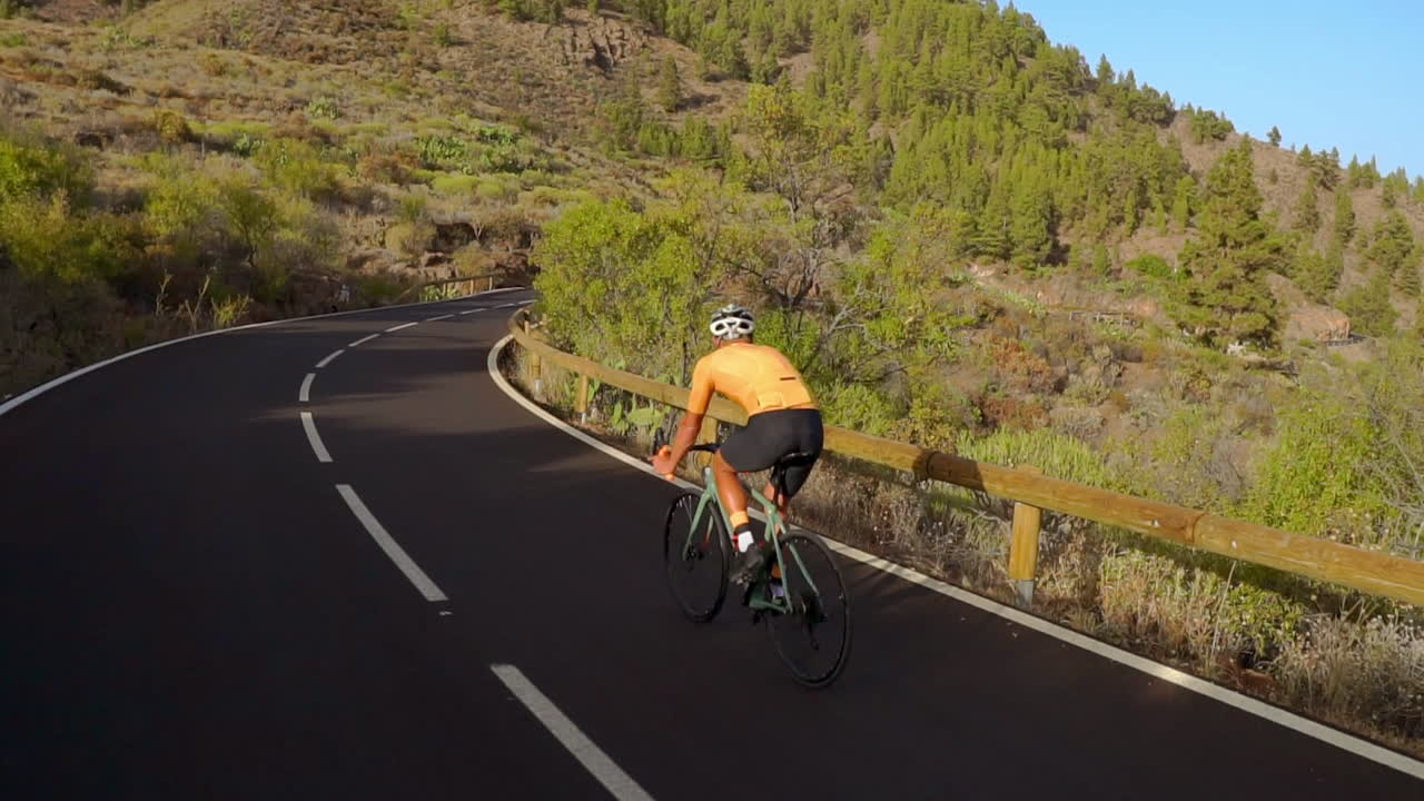 A tracking shot records a male cyclist's journey up a mountain road. He's undergoing cycling training on a sunny day along a hilly highway road