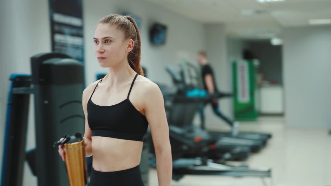 A slender woman in a black sports top and with her hair stuck in the tail is drinking water from a shaker and walking through the gym. Blurred Background