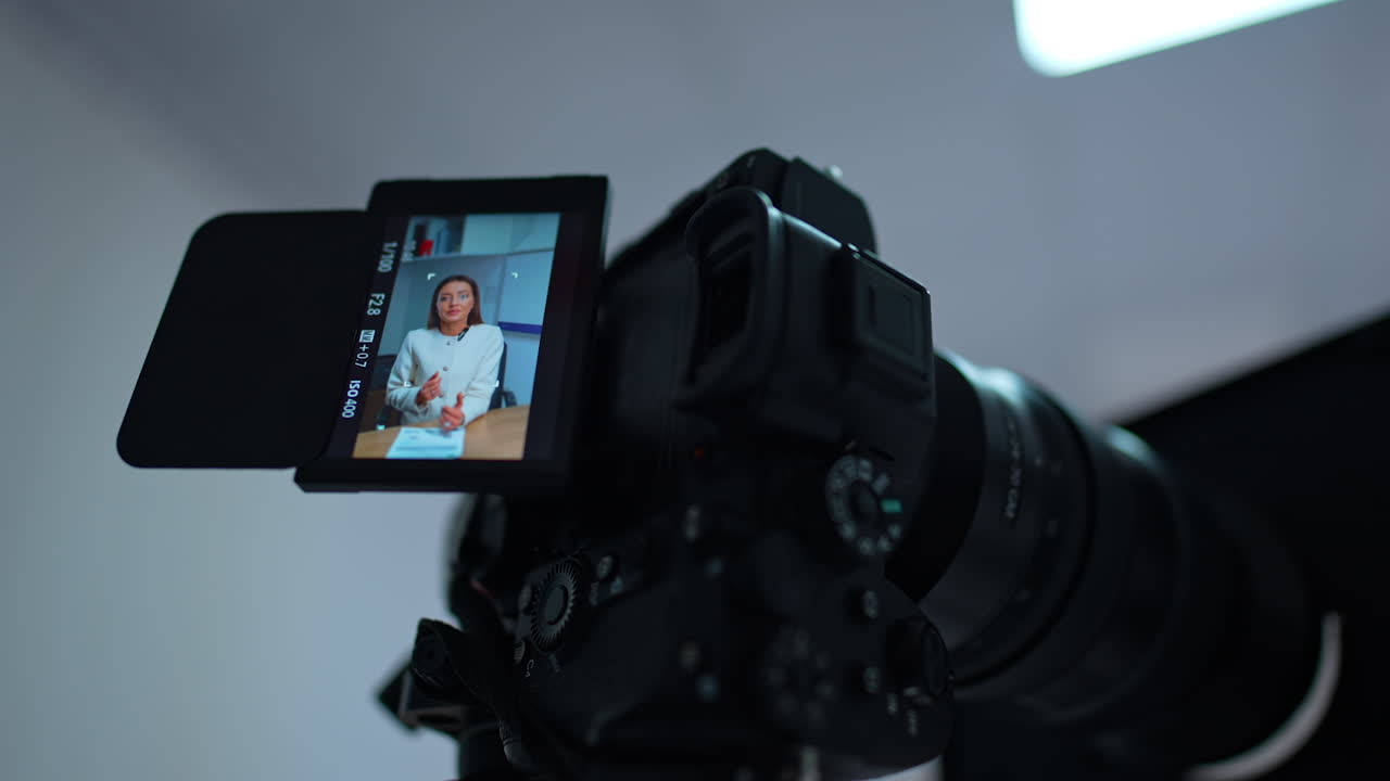 Caucasian woman with long dark hair wearing white jacket speaking on the camera display. Low angle view close up at the professional equipment for filming a blog content.