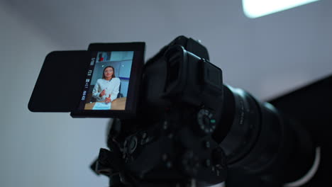 caucasian woman with long dark hair wearing white jacket speaking on the camera display. low angle view close up at the professional equipment for filming a blog content.