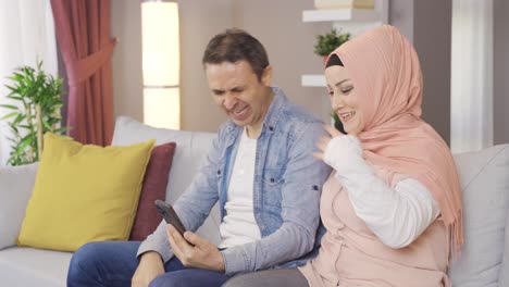 Muslimische-Frau-Im-Hijab-Und-Ihr-Mann-Schauen-Zu-Hause-Gemeinsam-Auf-Das-Telefon.