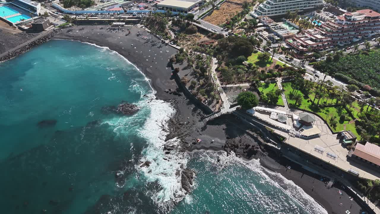 Vista aérea de la espectacular costa de Tenerife, con playa volcánica de arena negra, océano turquesa, acantilados y complejo turístico urbano