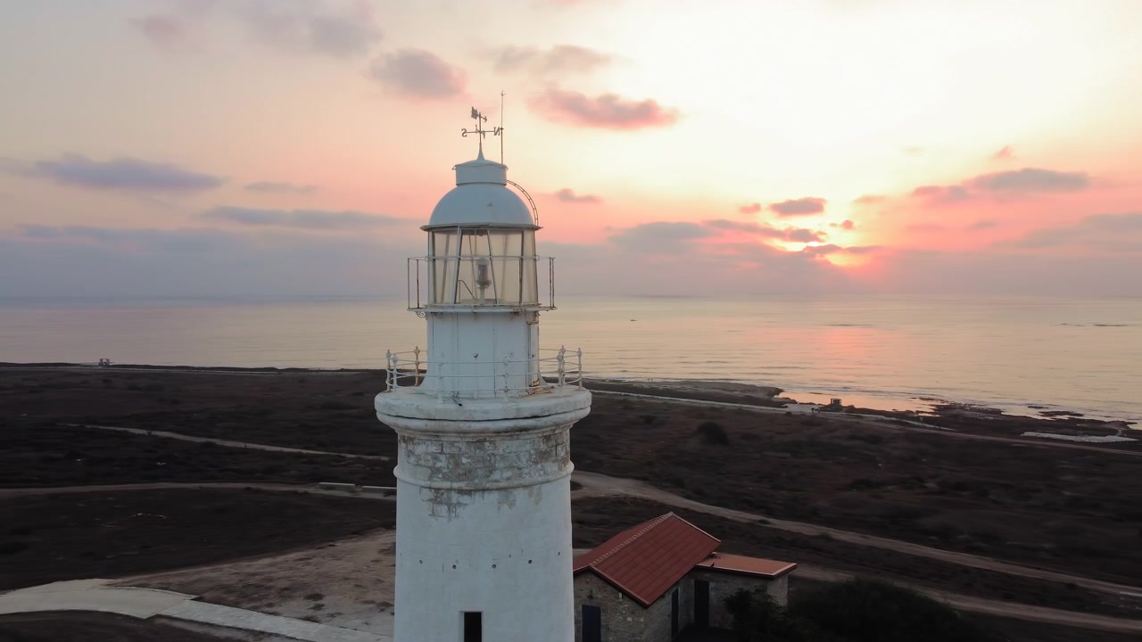 Un faro bianco si erge imponente contro un cielo pastello al tramonto, dominando l'oceano calmo e la costa.