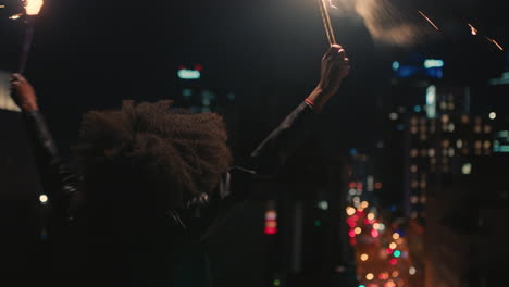 feliz joven afroamericana sosteniendo bengalas bailando en la azotea por la noche celebrando la víspera de año nuevo disfrutando de la celebración navideña