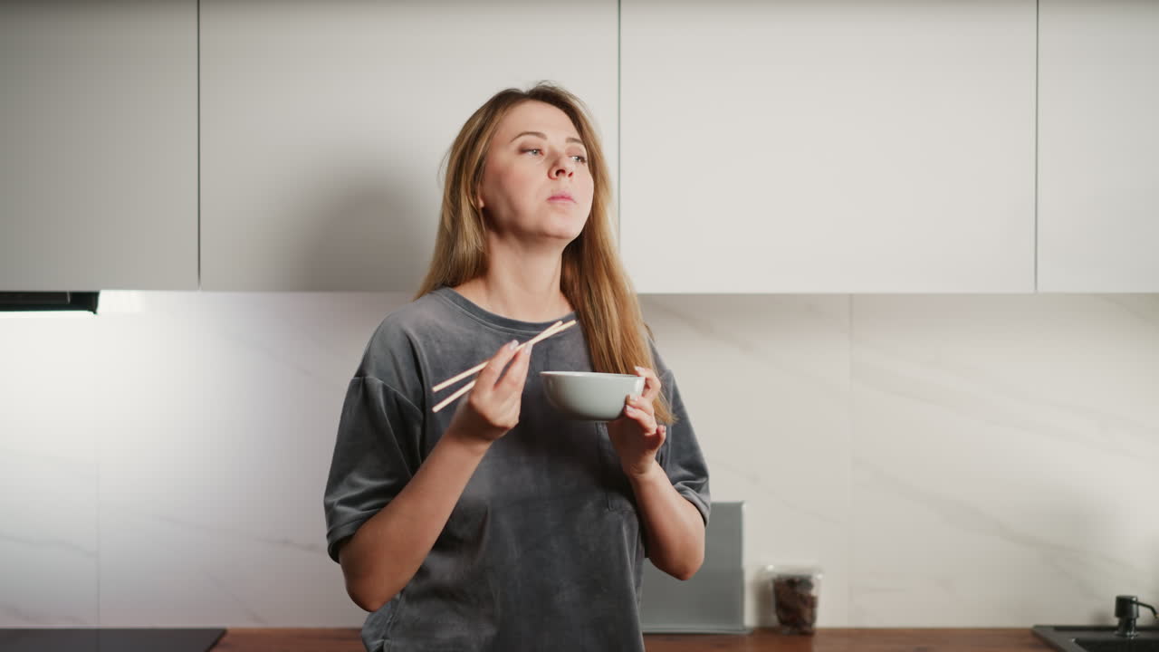 Hungry young girl in oversized t-shirt standing in modern kitchen enjoying spaghetti with chopsticks, savoring quick homemade meal in relaxed home