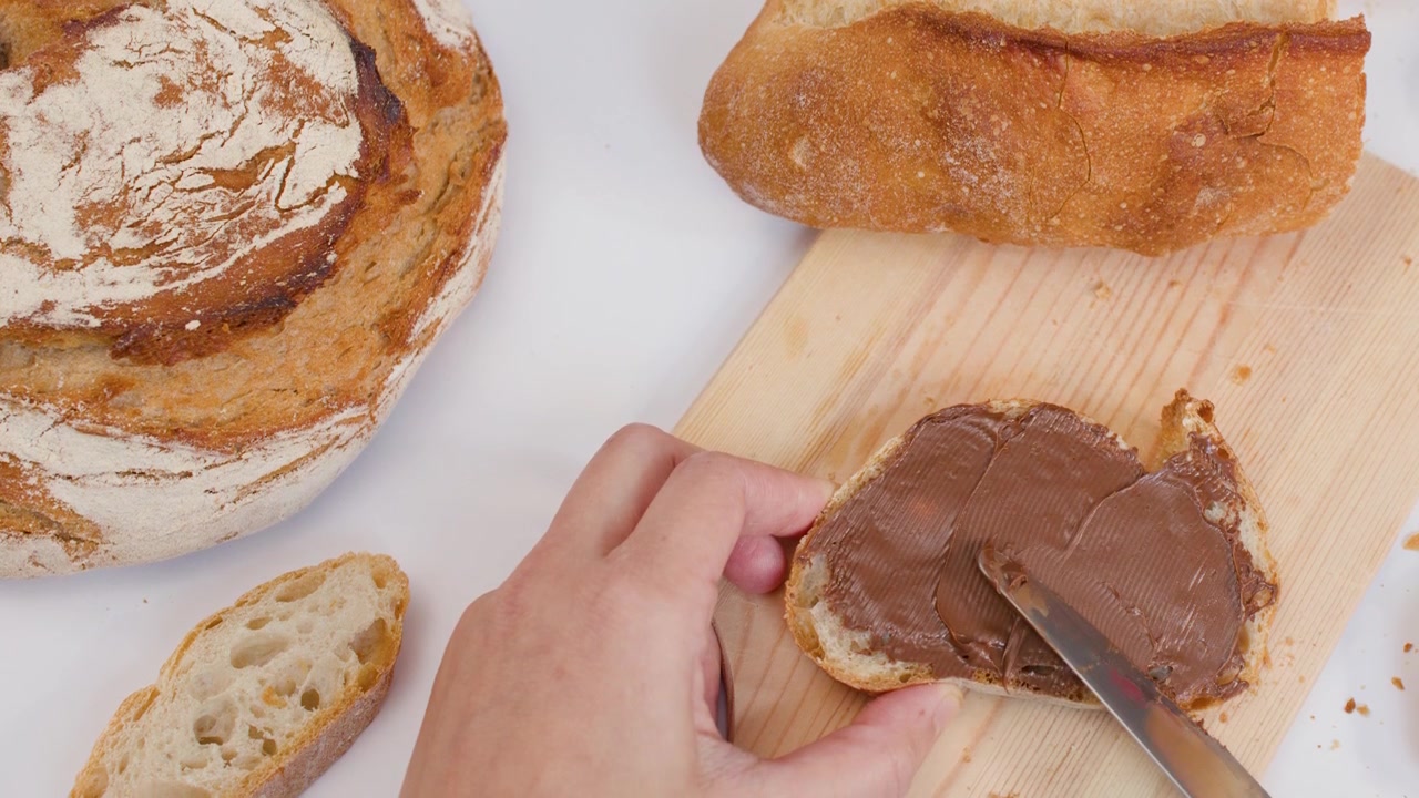Hand spreads chocolate cream on sourdough bread over wooden board against white background.