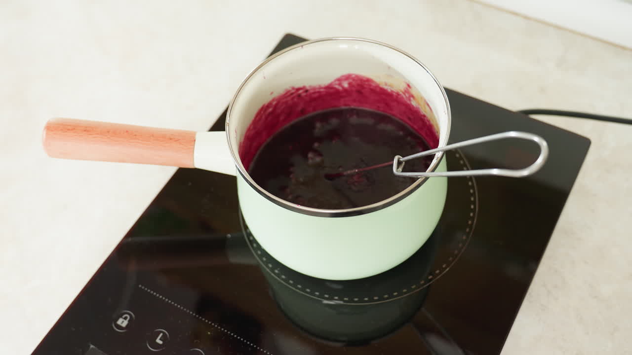 Close up of red soup or sauce simmering inside light green pot on black electric stove with visible mixing utensil, showcasing cooking process and heat effect on thick, vibrant liquid preparation
