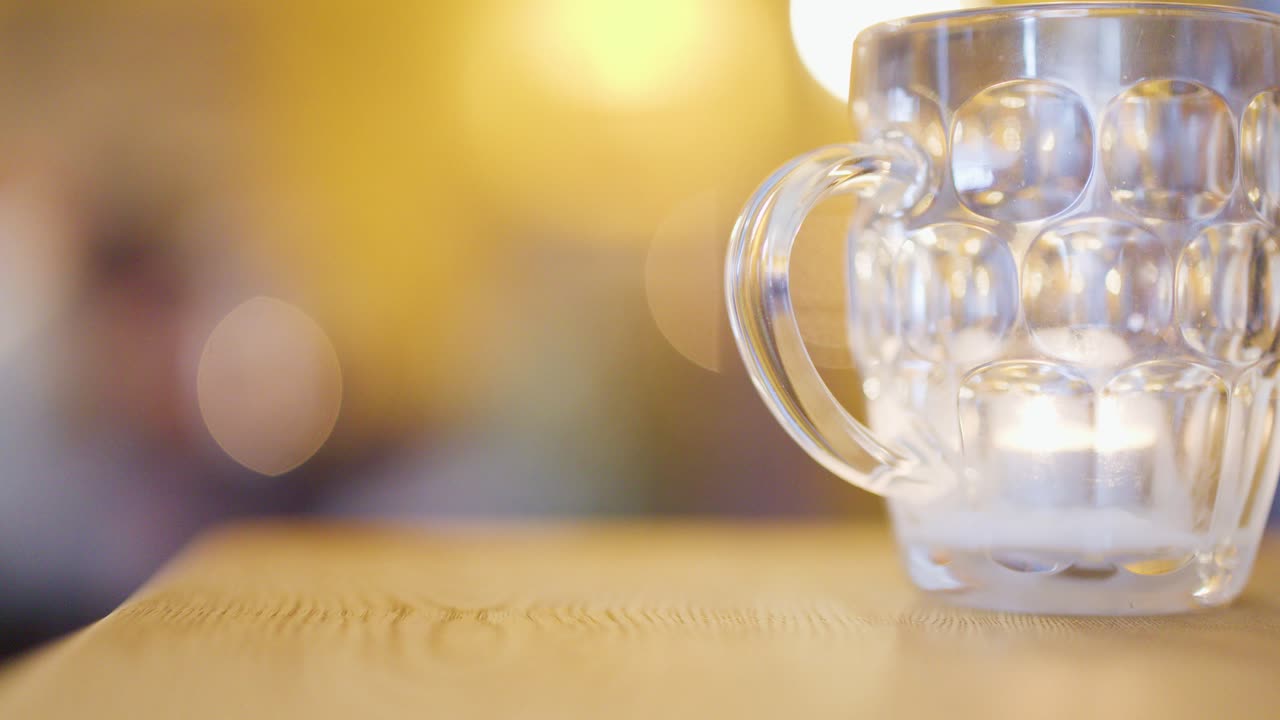 A clear glass mug with a lit candle sits on a wooden table, surrounded by warm, ambient bokeh lighting and a softly blurred background