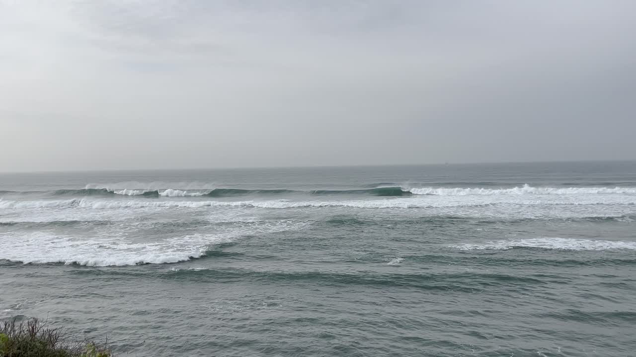 Gentle waves roll across the calm sea along the Portuguese coastline, creating a peaceful and expansive view of the beach and horizon under a soft, overcast sky.