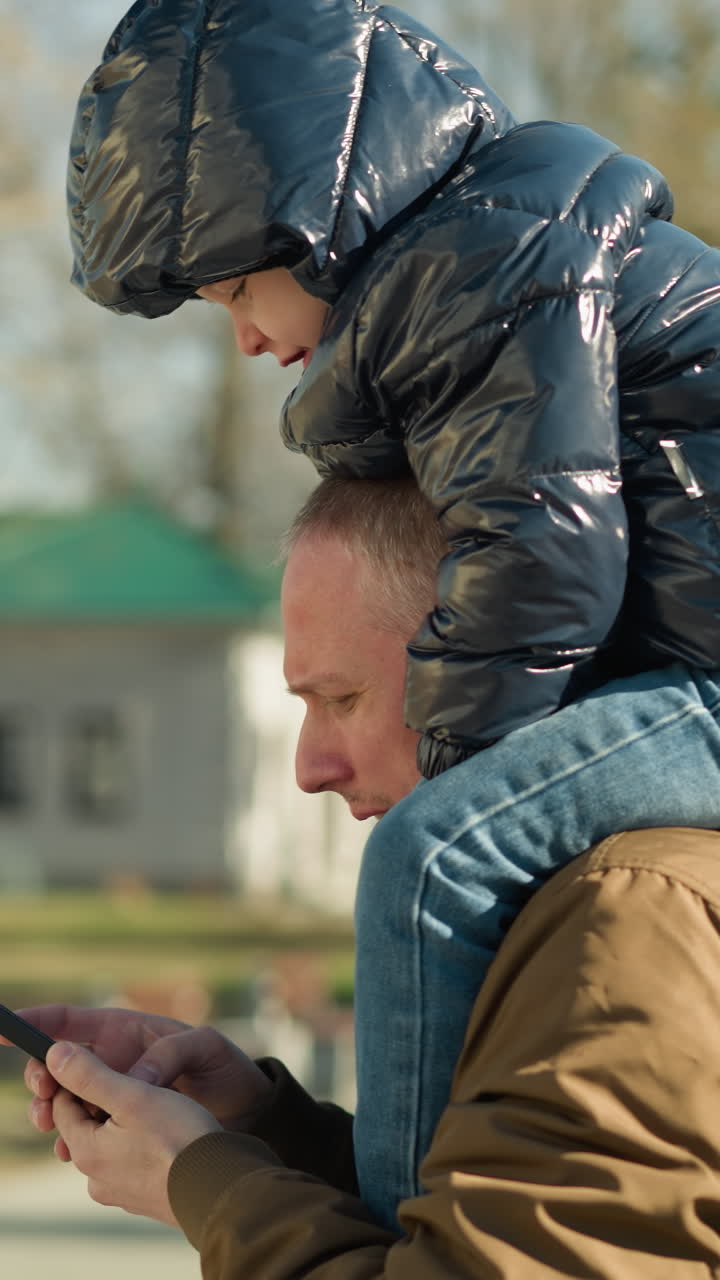 primer plano un padre llevando a su hijo en sus hombros mientras opera su teléfono girado hacia el lado derecho, con algunos caminando hacia abajo, con árboles y edificios detrás