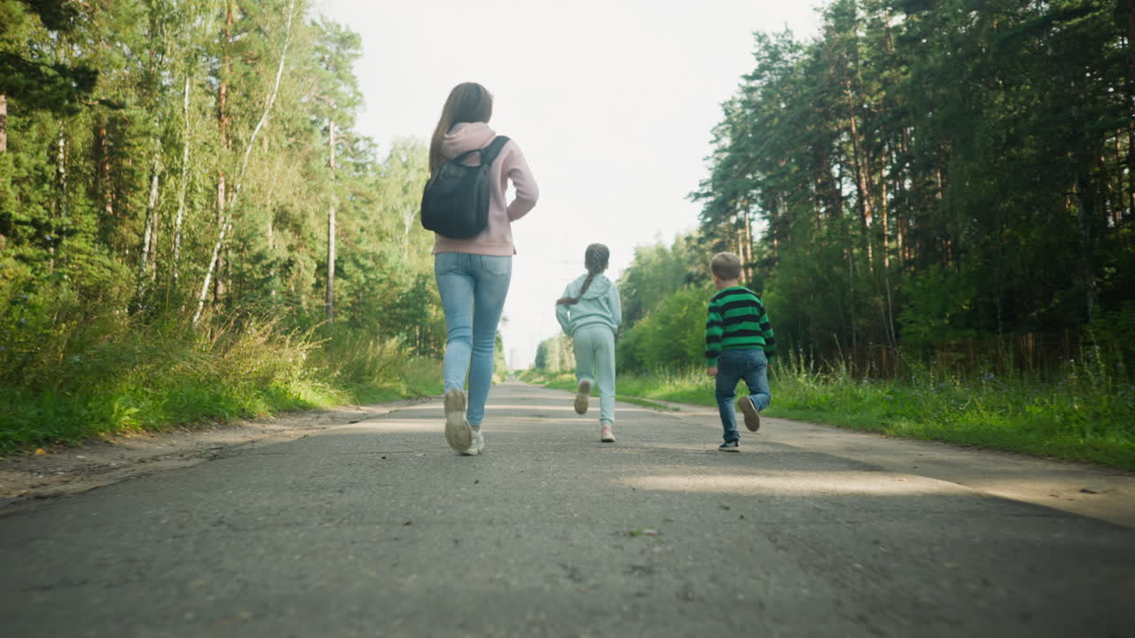 Rear view of cheerful family moment as young woman and two children run joyfully on paved rural road surrounded by tall green trees, creating a playful, bonding scene under soft natural daylight