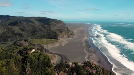 aerial footage drone close up to cliff mountain rock formation reveal lonely endless black sand beach with blue ocean big wave on comans track, karekare, new zealand