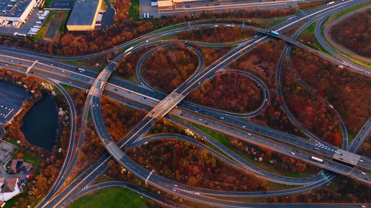 Cloverleaf highway interchange surrounded by autumn woods, New Jersey. Top-down of looping cloverleaf ramps and an overpass weaving through colorful seasonal trees and warehouses