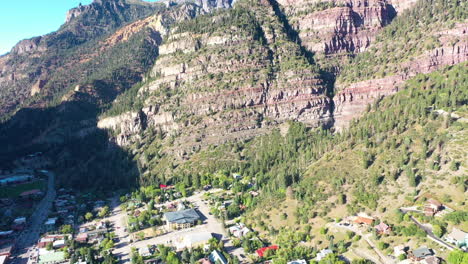 aerial drone tilt down of beautiful ouray colorado mountain town surrounded by thick pine tree forest and cliffs during summer with houses and cars driving