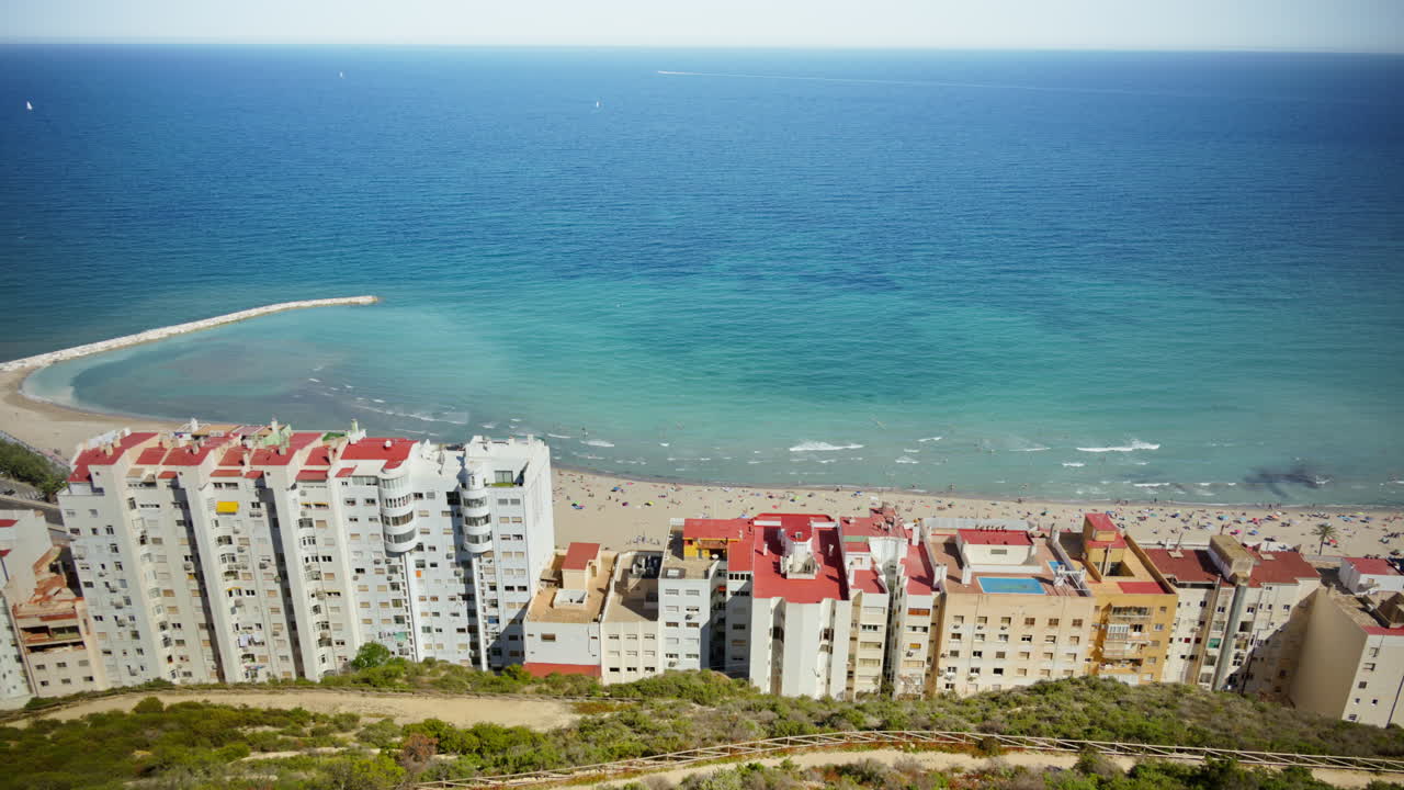 Aerial view of turquoise water, a sandy beach and seafront apartment blocks with swimmers and umbrellas visible below