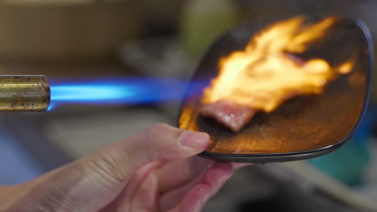 A seared beef aburi sushi being blow torched with a flame on a black plate in a Japanese restaurant