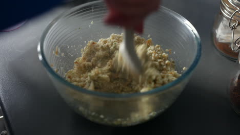a hand stirring stuffing mix in a glass bowl, blending ingredients into a textured, moist consistency. the partially mixed stuffing sits on a countertop near jars of seasonings, ready for cooking