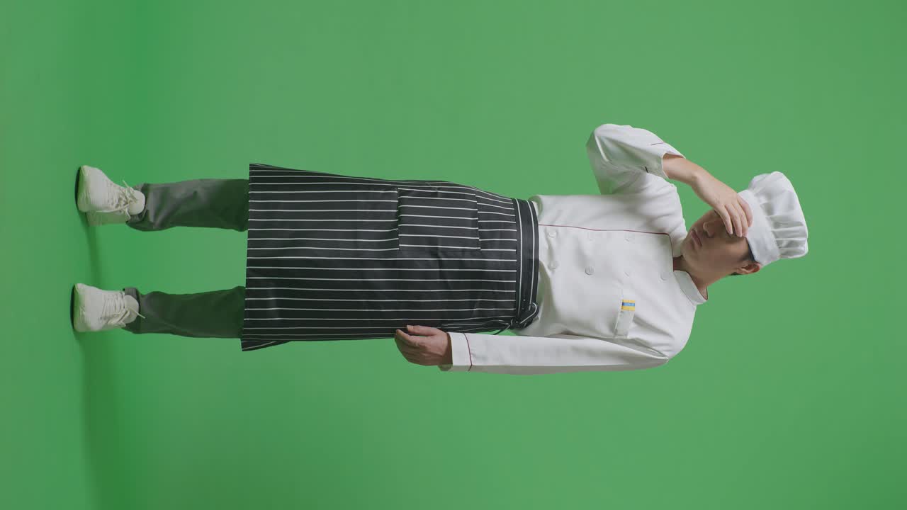 녹색 스크린 배경 스튜디오에 서 있는 동안 두통을 가진 아시아 남성 요리사의 몸이 가득 차 있습니다.