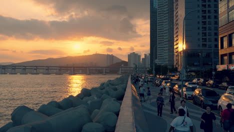 silhouetted korean people walking at busan marina bay broadwalk at sunset with a view of gwangan bridge, tourists enjoy watching sun setting over mountain, sunlight reflections in sea and building