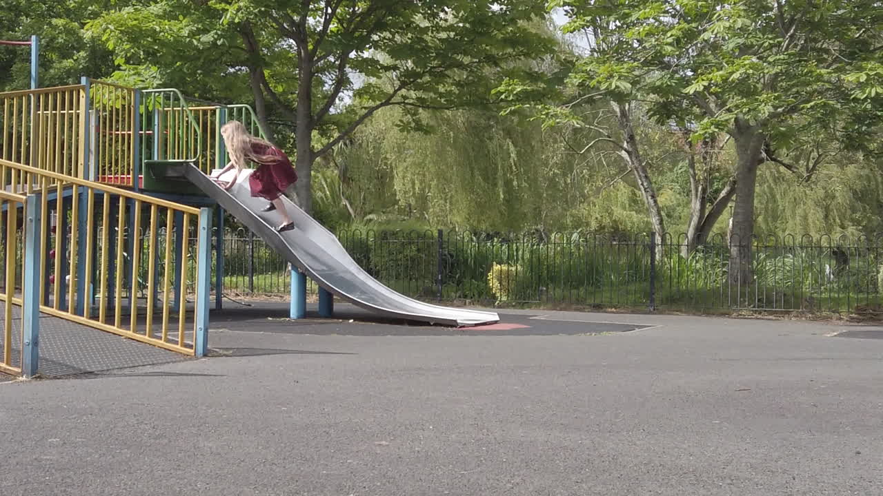 A static wide shot captures a child standing on the metal playground slide in Victoria Park, Paignton, UK. The shot emphasizes the architectural lines of the playground structure against the park's gr