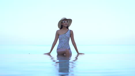 mujer joven con clase en traje de baño, sombrero y gafas de sol sentada en el borde de la piscina infinita con un interminable horizonte marino tropical en el fondo, fotograma completo