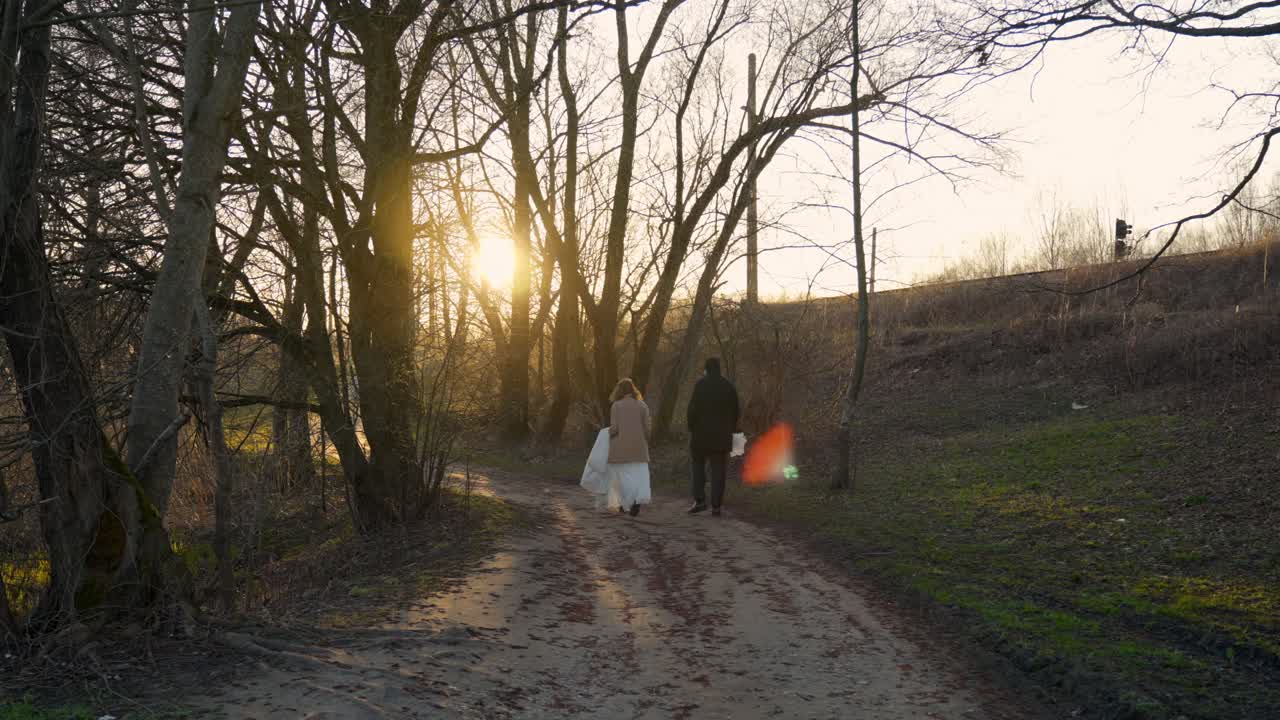 A romantic wedding couple walking on a forest path at sunset, captured from behind with sunlight.