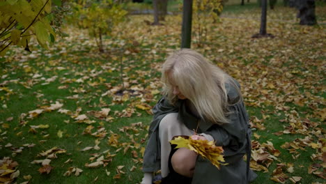 zoom shot of beautiful senior woman in grey coat collecting leaves in the park.